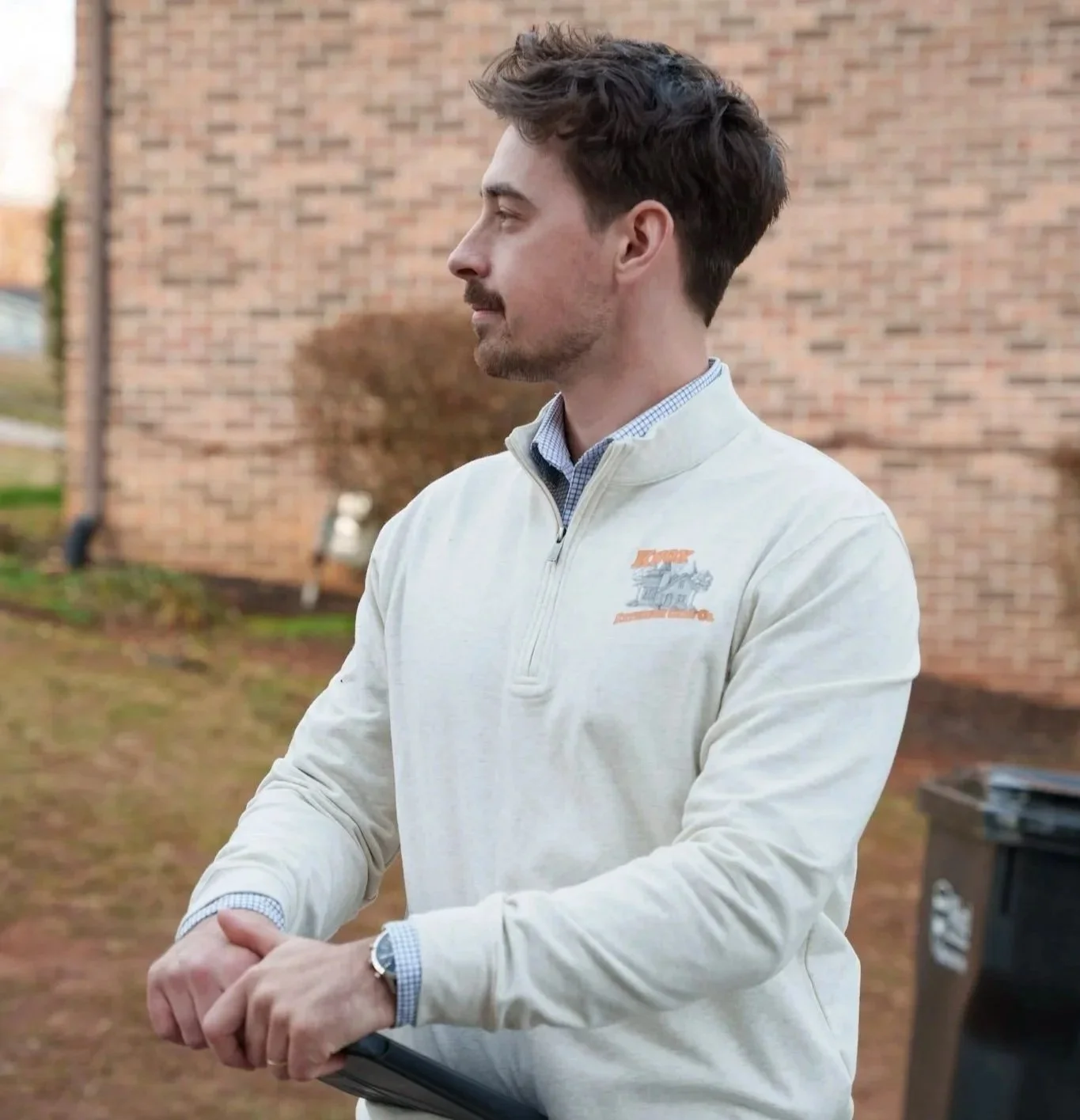 A man standing outdoors in profile, wearing a light-colored quarter-zip sweater with a logo on the chest, and holding a clipboard, with a brick building and a trash bin in the background.
