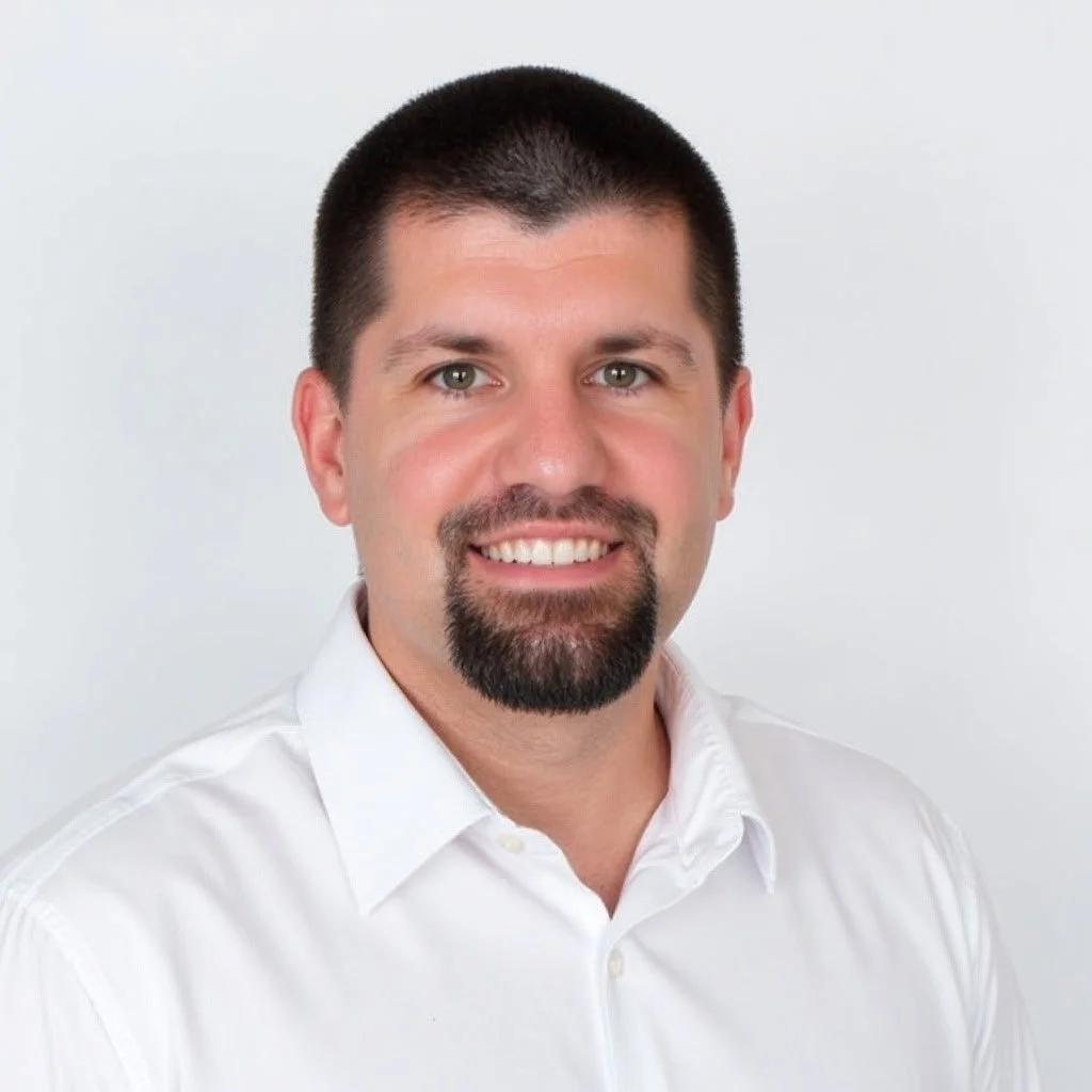 A man with short dark hair, a goatee, and a white shirt, smiling at the camera with a plain white background.