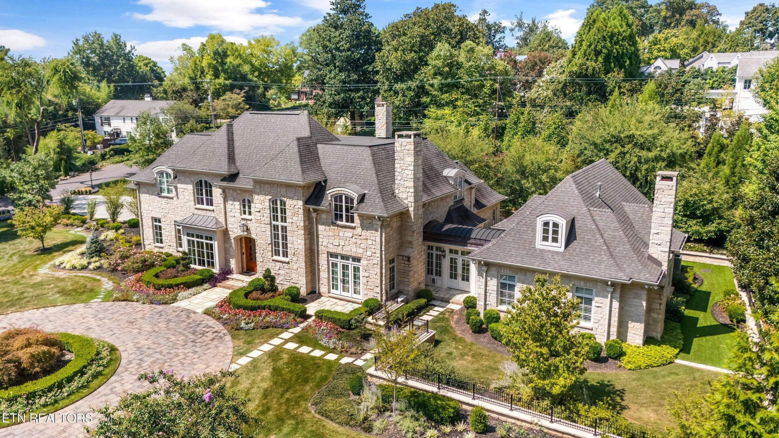 A large, elegant house with stone exterior walls, surrounded by lush landscaping, trees, and a well-manicured lawn, with a curved stone driveway in the front.