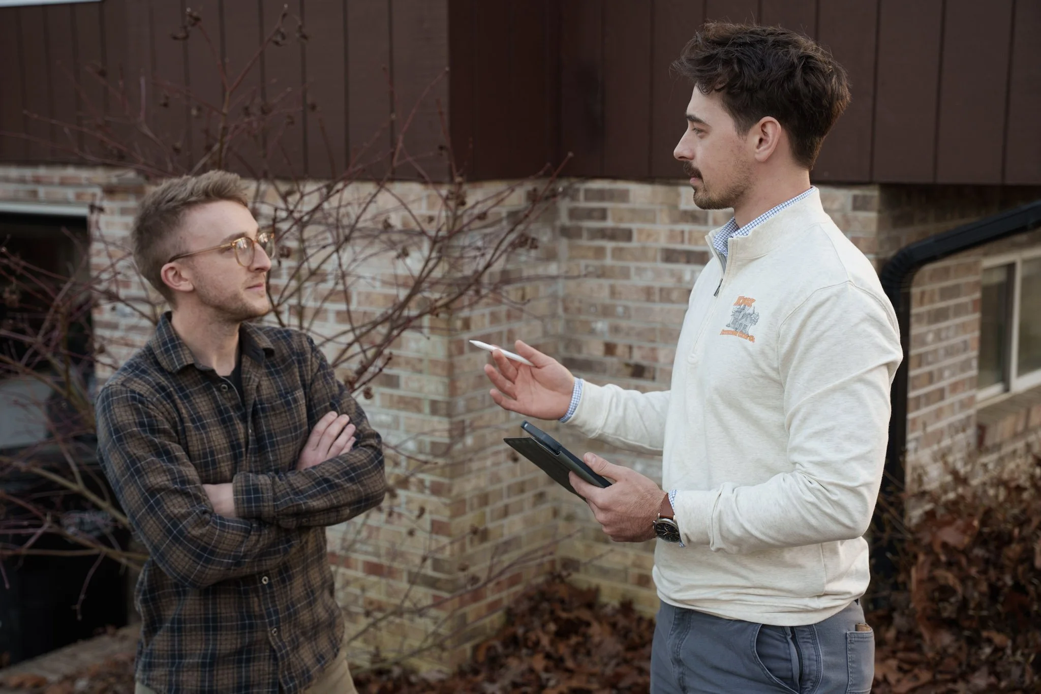 Two men having a conversation outside near a brick house. One man is wearing glasses and a checkered shirt, with arms crossed. The other man, holding a tablet and a small object, is wearing a light-colored zip-up jacket and a watch.