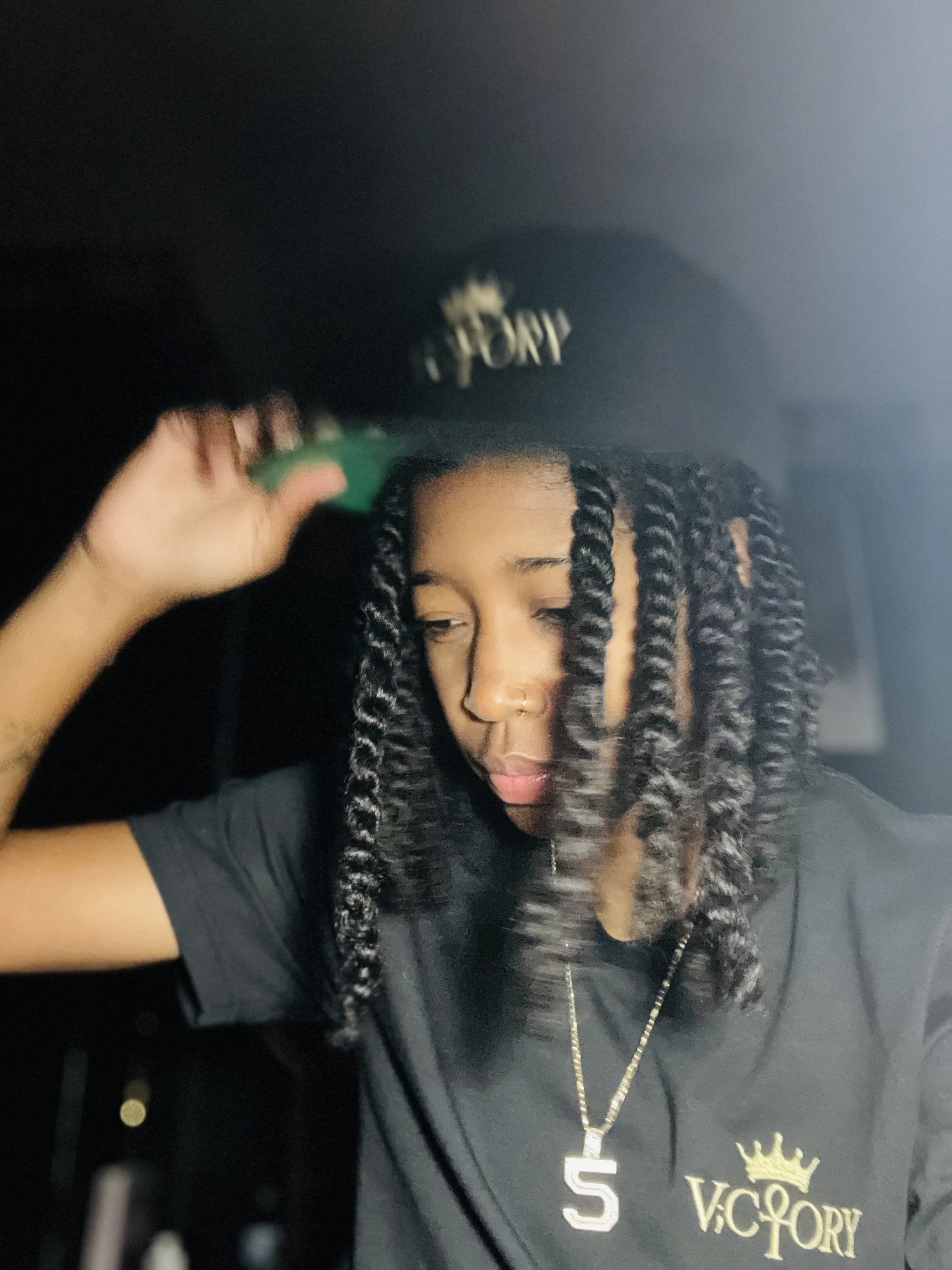 Person with medium skin tone and black twisted hair wearing a black V.C. Gregory cap and black T-shirt with gold logo, adjusting the cap in a dimly lit environment.