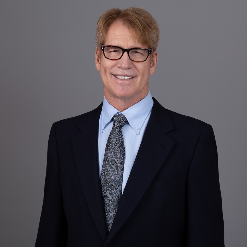 A middle-aged man with light brown hair, wearing black-rimmed glasses, a black suit, a light blue dress shirt, and a patterned tie, smiling against a gray background.