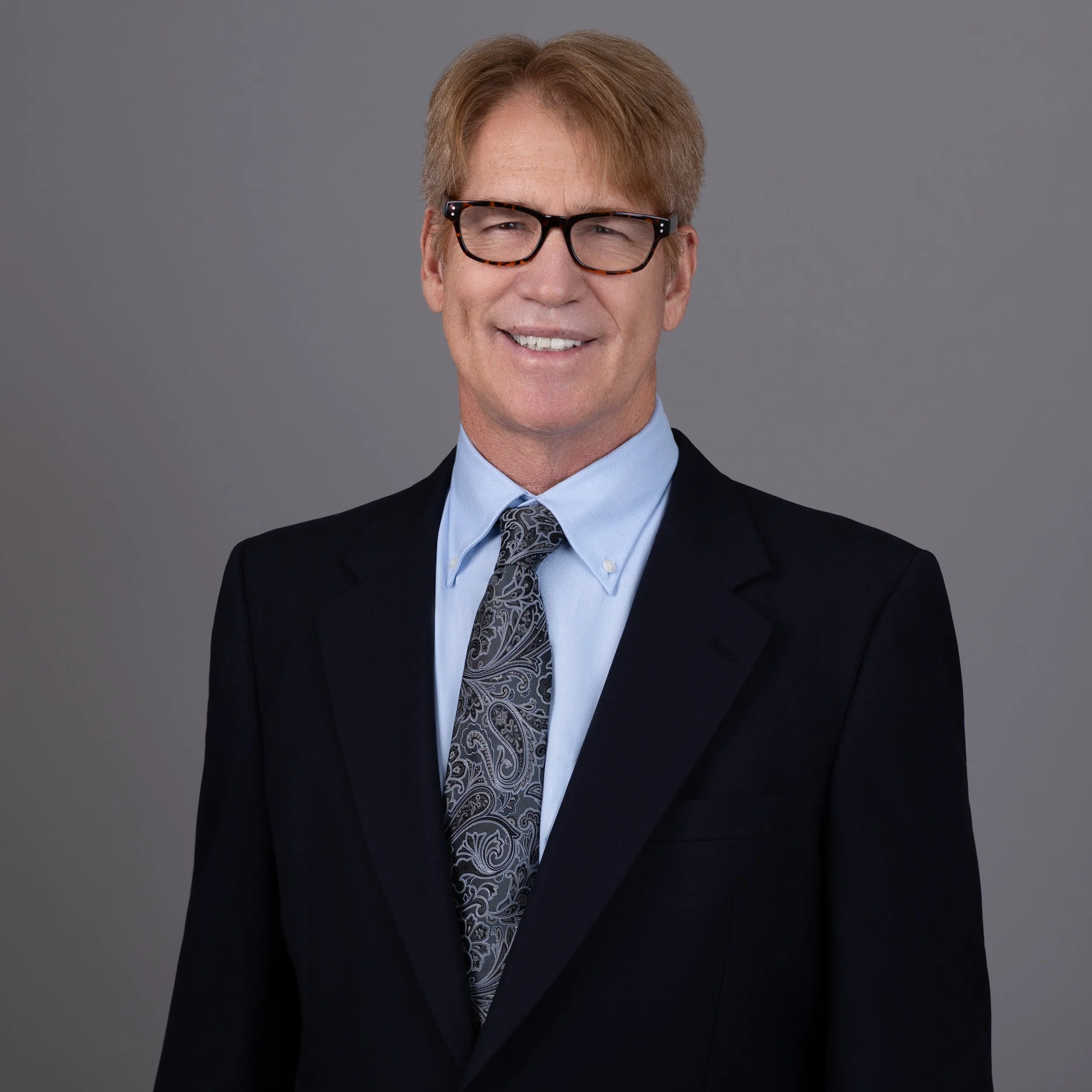 Headshot of a middle-aged man with reddish-brown hair, wearing black-framed glasses, a dark suit, light blue dress shirt, and a patterned tie, smiling against a gray background.