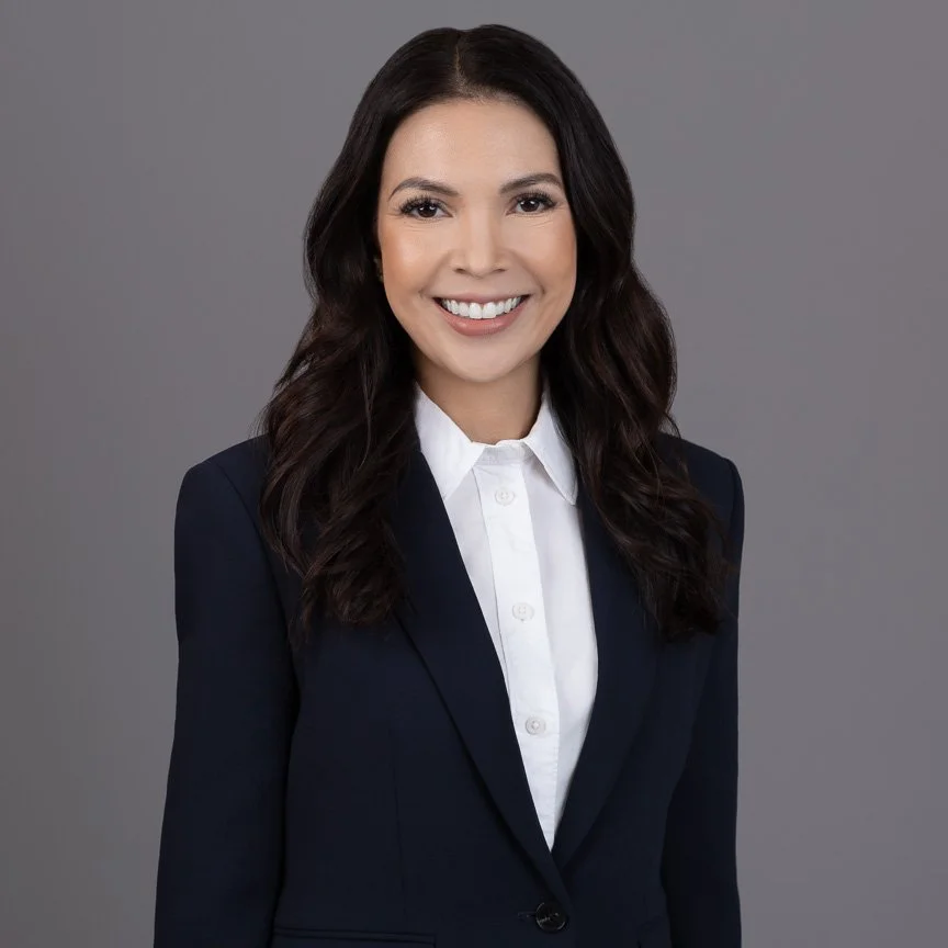 A professional woman with long dark hair, wearing a navy blazer and white shirt, smiling against a gray background.
