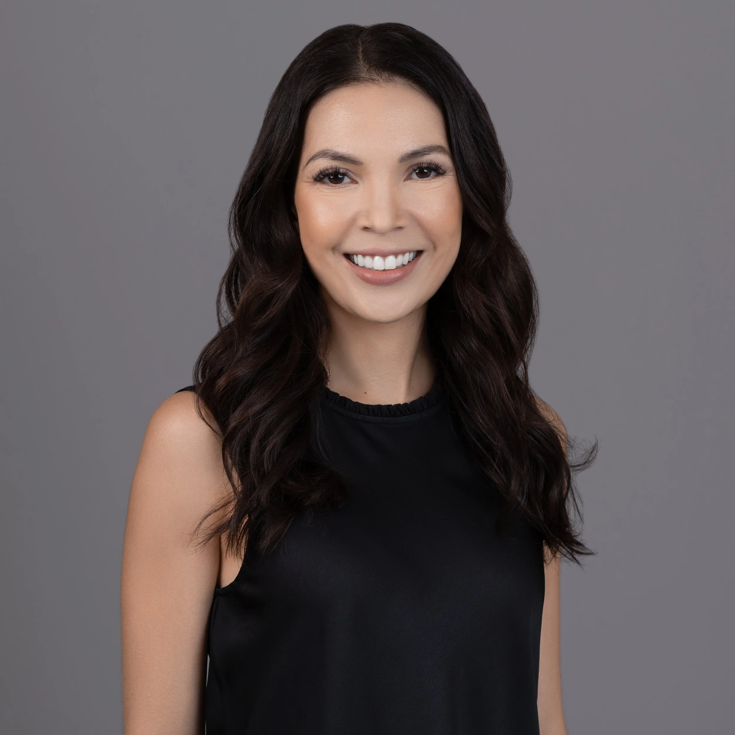 Close-up of a woman with long dark hair, smiling, wearing a black sleeveless top, against a gray background.