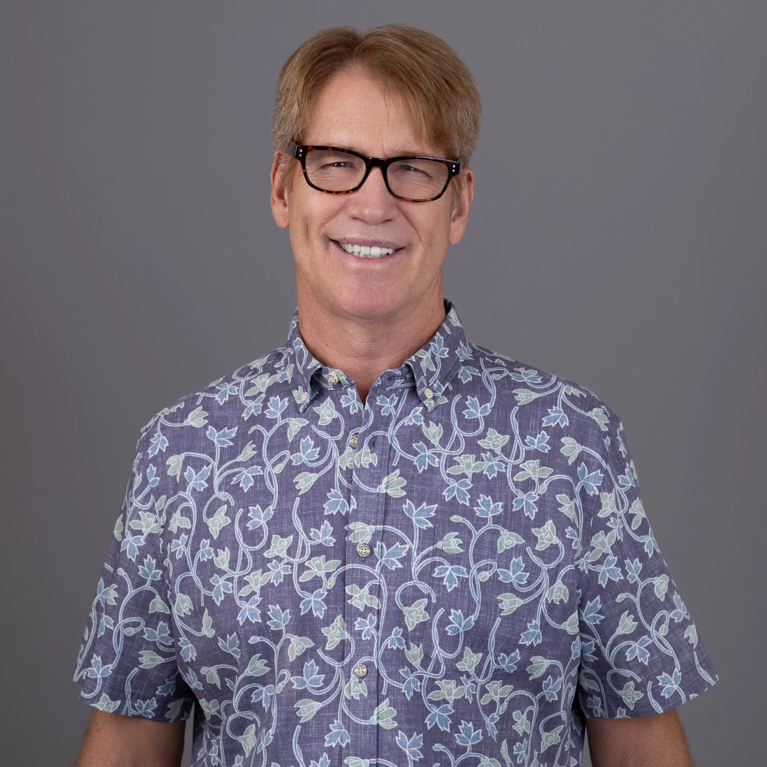Close-up of a smiling man with light skin, reddish-blonde hair, black framed glasses, and wearing a purple shirt with a floral pattern.