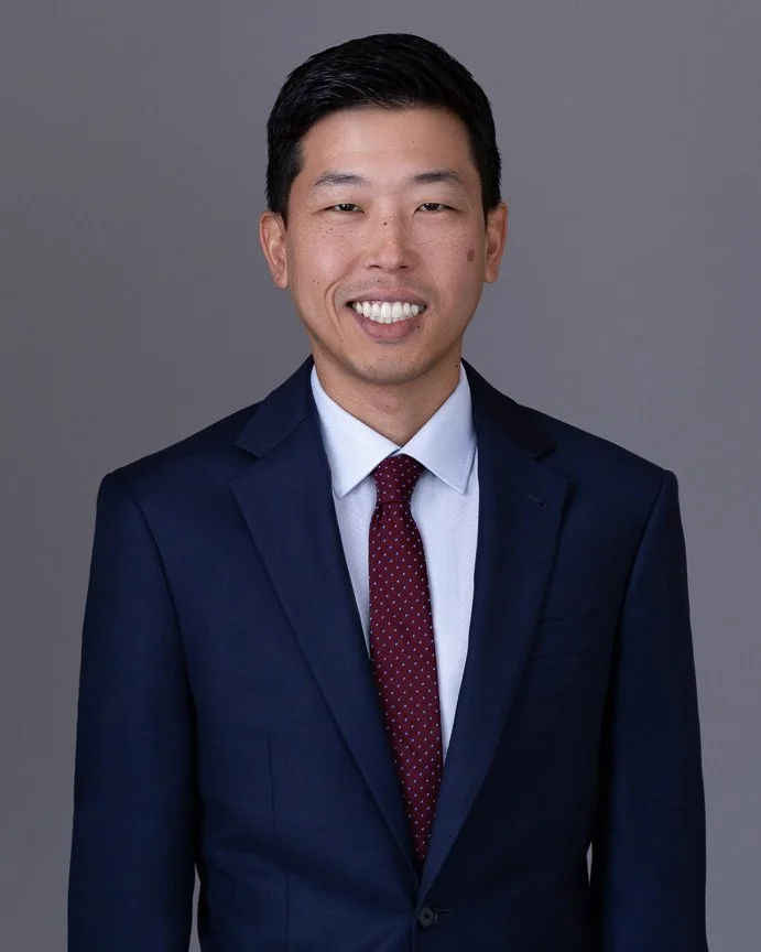 A man in a dark blue suit, white dress shirt, and maroon polka dot tie smiling against a gray background.