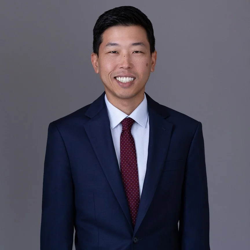 Portrait of a young Asian man in a navy blue suit, white shirt, and red tie, smiling against a gray background.