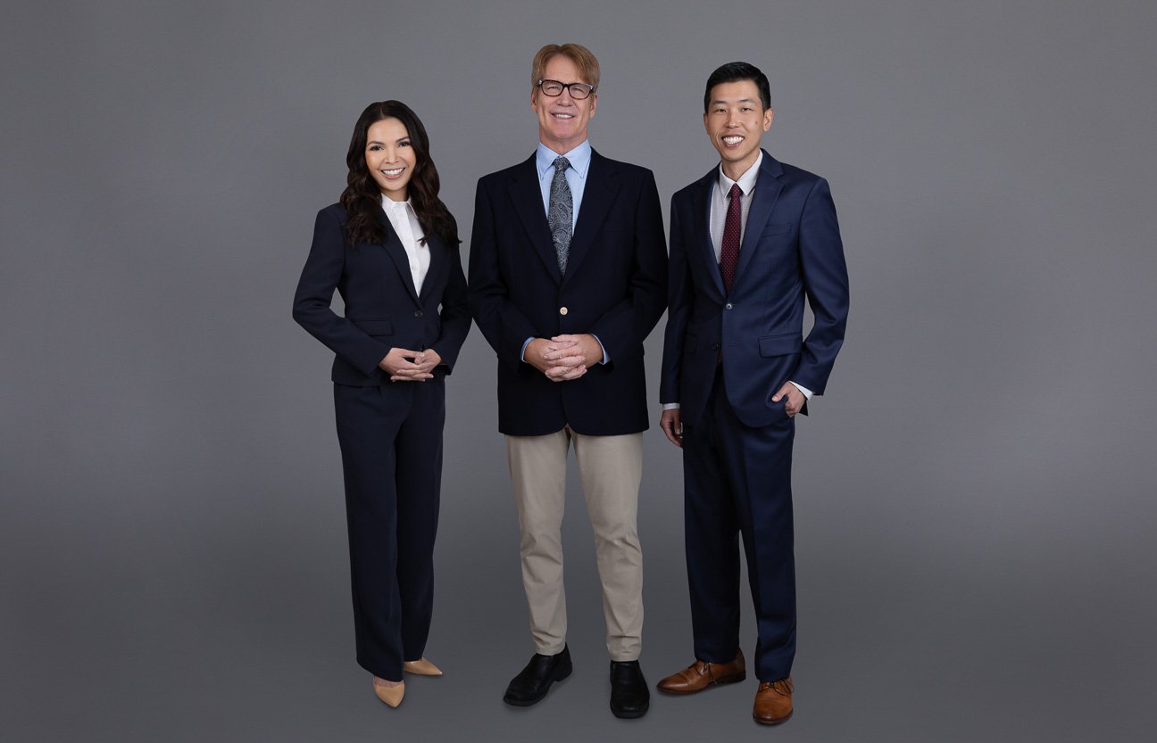 Three professionally dressed individuals standing together against a gray background, smiling and looking at the camera.