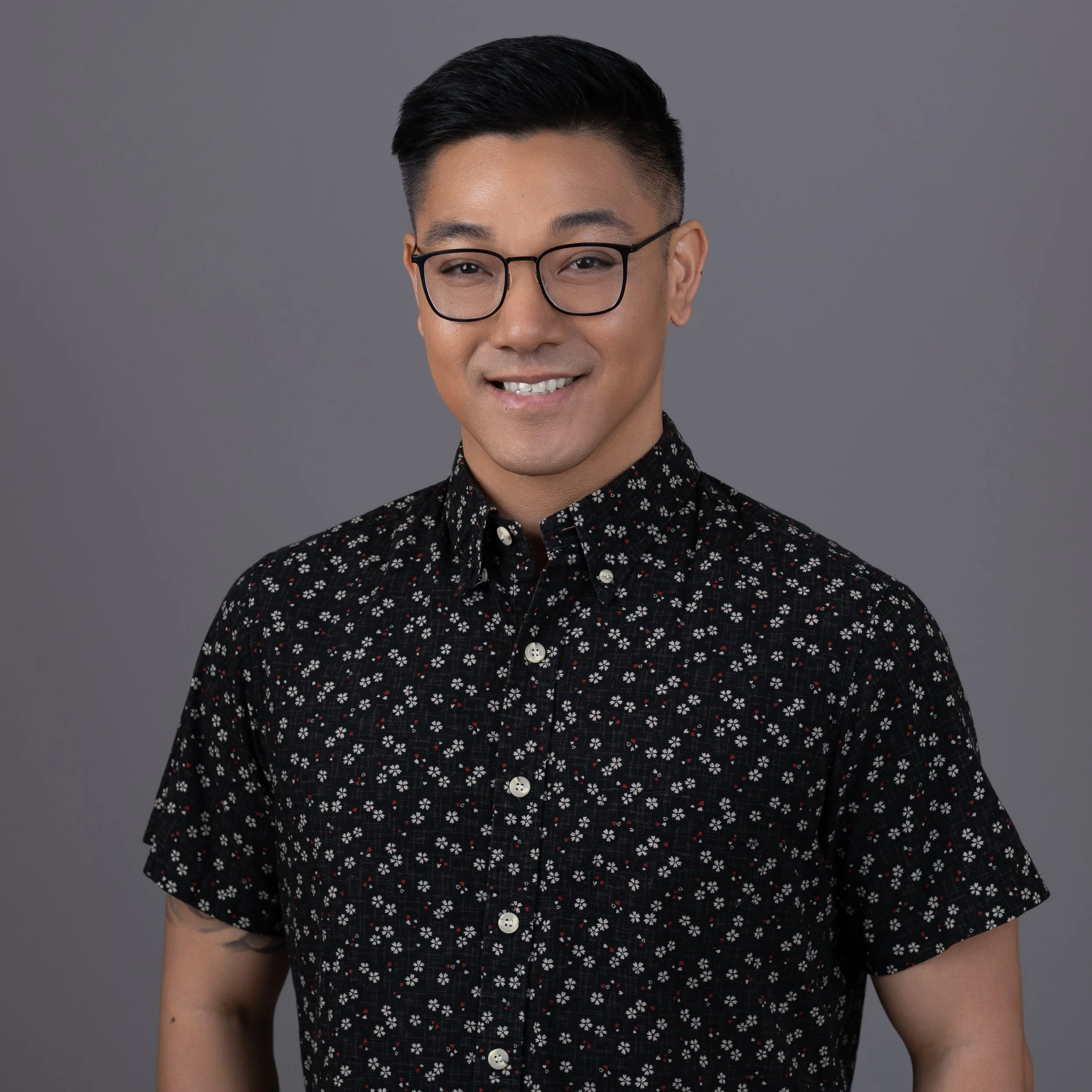 Close-up of a person wearing glasses and a black shirt with white and red floral pattern, smiling against a gray background.
