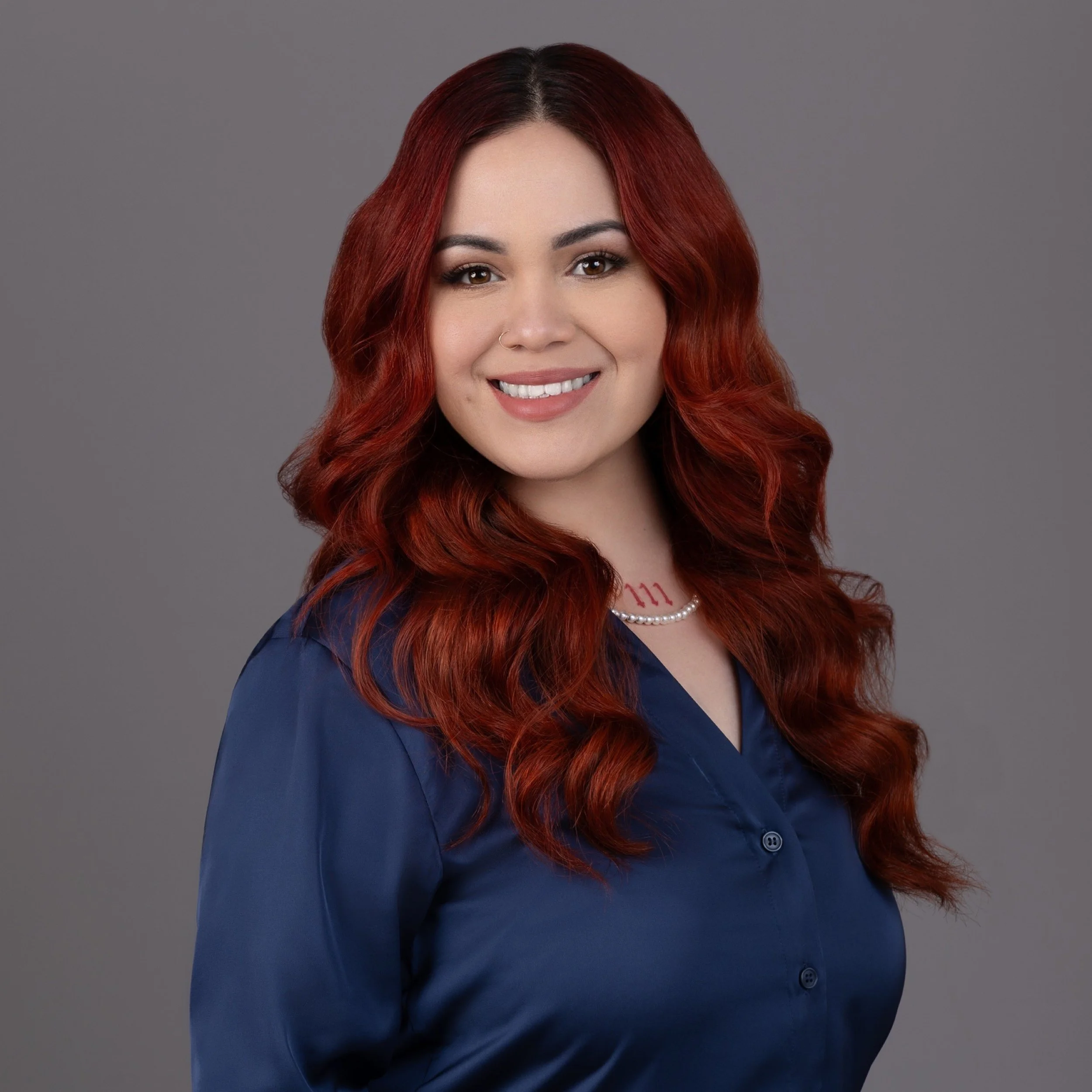 A woman with red, wavy hair smiling against a gray background, wearing a dark blue blazer, a pearl necklace, and a nose ring.