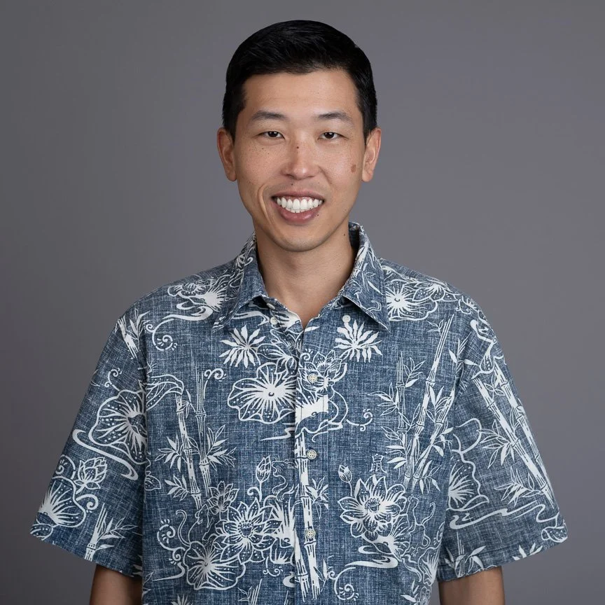 A young Asian man smiling, wearing a blue Hawaiian shirt with white floral and leaf patterns, against a gray background.