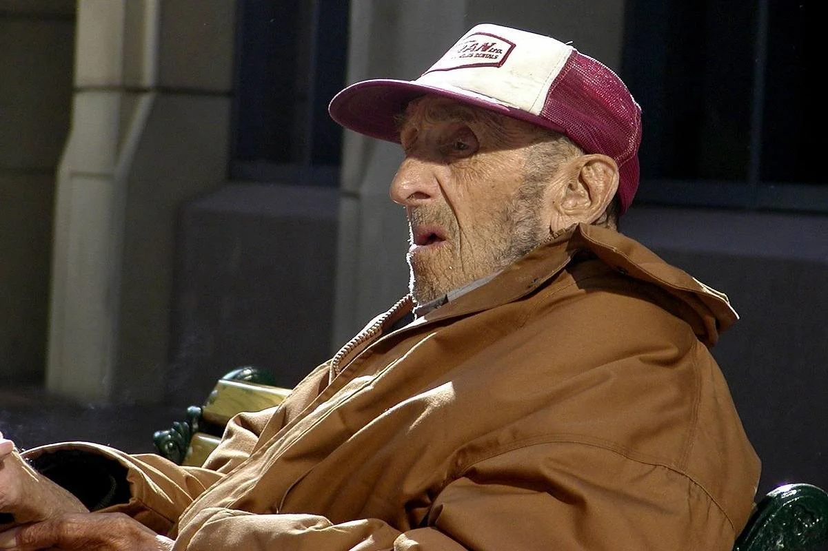 An elderly man wearing a brown jacket and a red and white trucker hat, sitting and looking to his right with a serious expression.