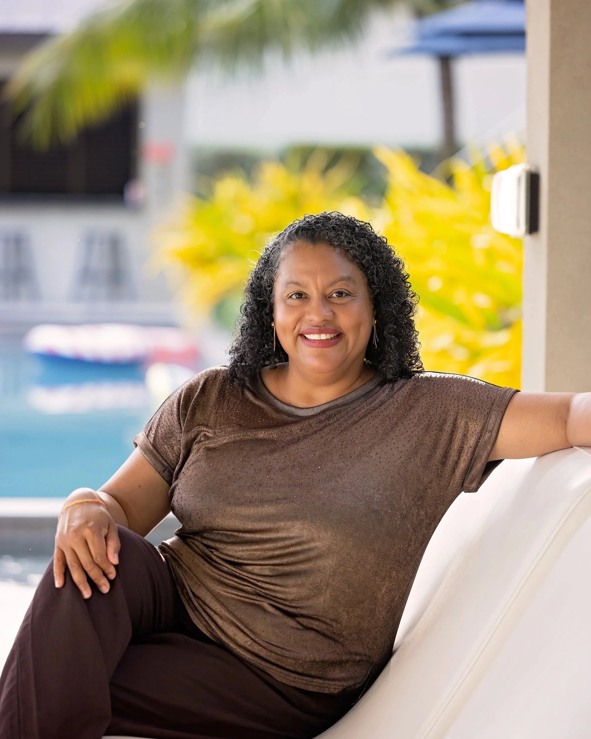 A woman with dark curly hair smiling while sitting on an outdoor white bench with a blurred background of plants and buildings.