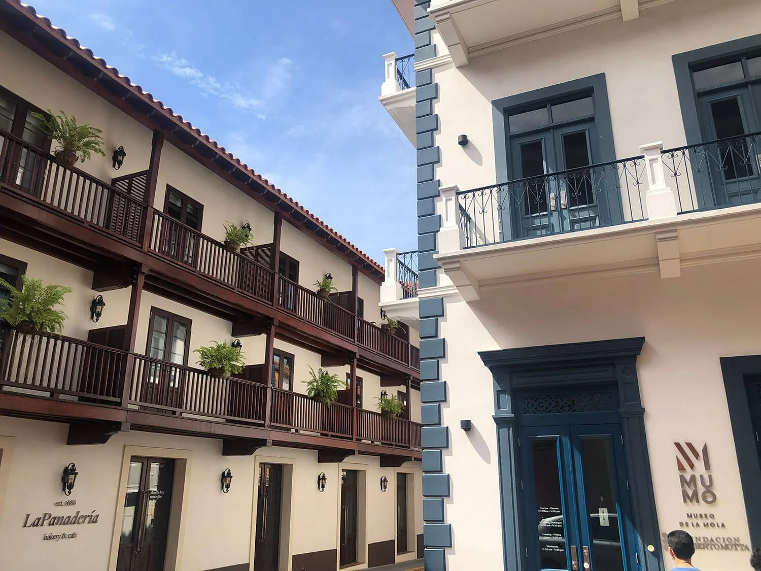 Photo of two adjacent buildings with contrasting architectural styles. The left building has a traditional style with brown wooden accents and multiple balconies with potted plants. The right building has a modern style with white walls, blue accents