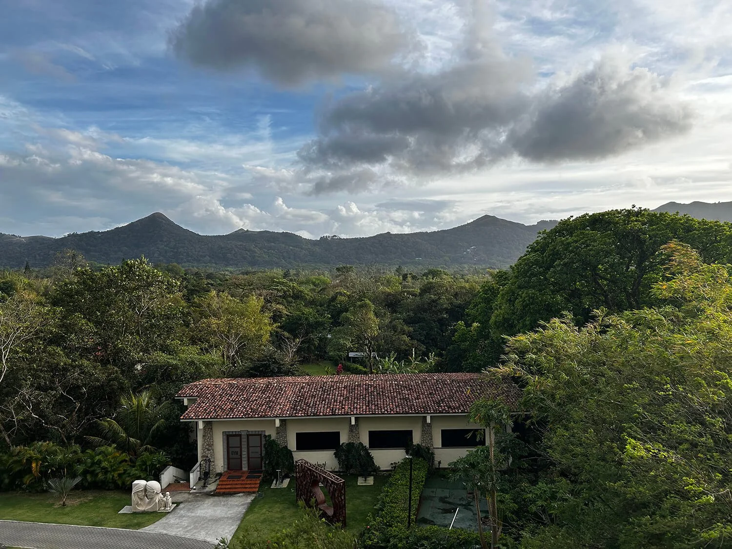 A house with a red-tile roof surrounded by lush green trees and mountains in the background under a partly cloudy sky.