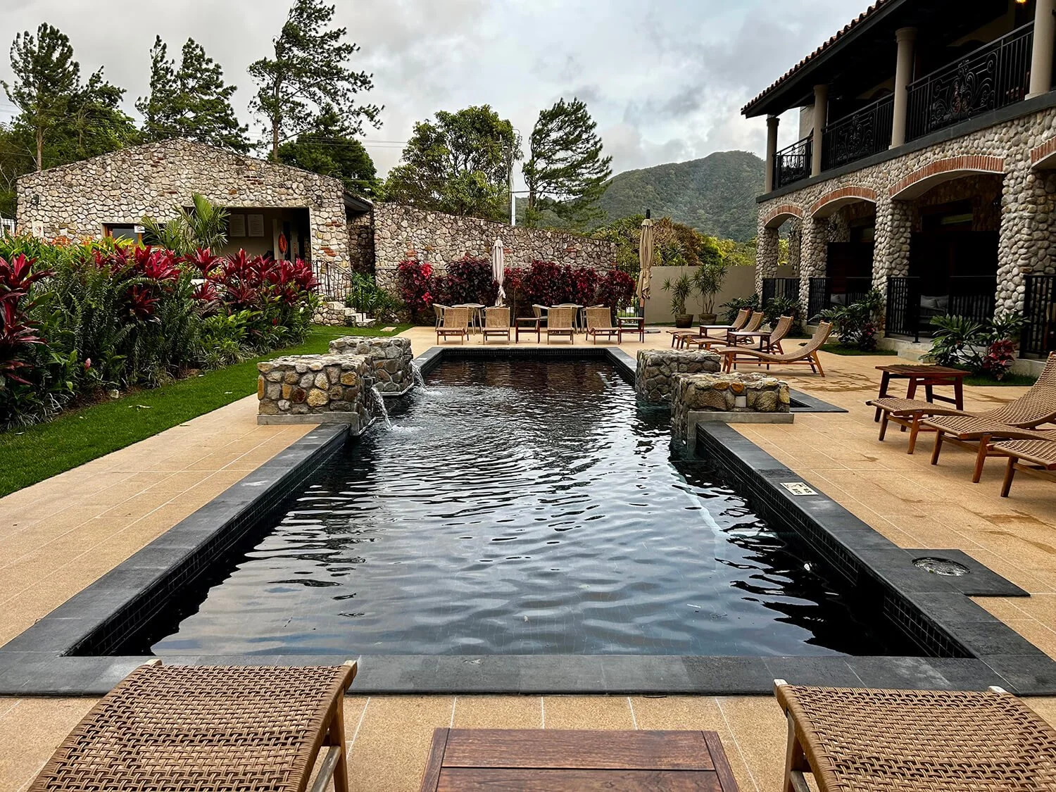 Outdoor pool area with lounge chairs, colorful plants, stone buildings, and mountain view.