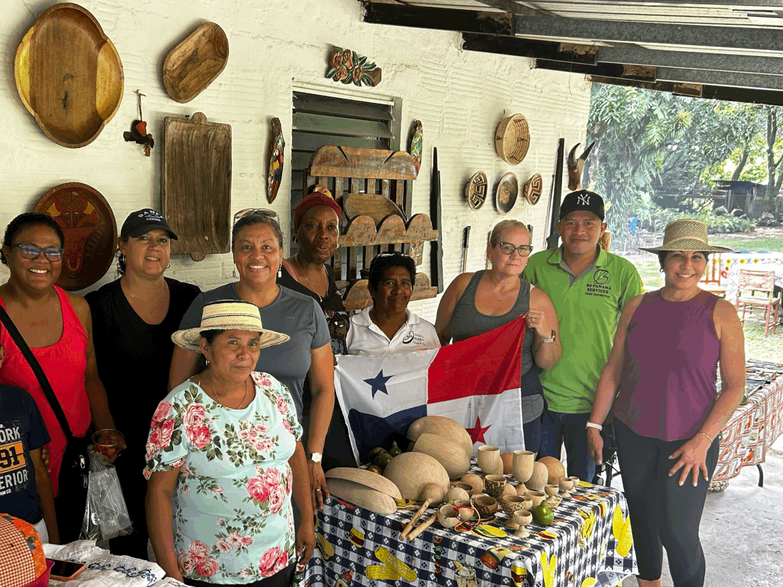 Group of diverse women and one man standing behind a table displaying handmade wooden crafts, with a woman holding a Panamanian flag, inside a decorated room with various wall hangings.