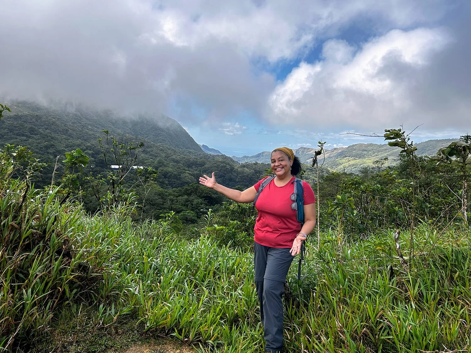 Woman in red shirt and gray pants standing in lush green mountainous landscape, smiling and gesturing towards the distant mountains under partly cloudy sky.
