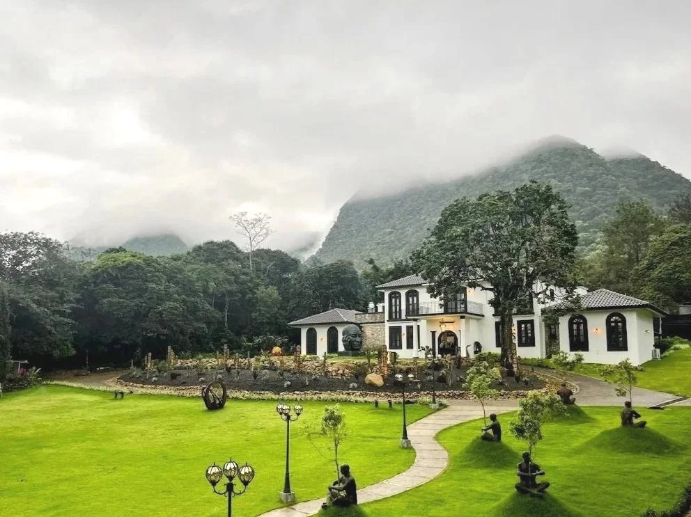 A large white house with black trim situated on a lush green lawn with elderly people sitting on benches. The house is surrounded by trees, with mountains in the background and a cloudy sky overhead.