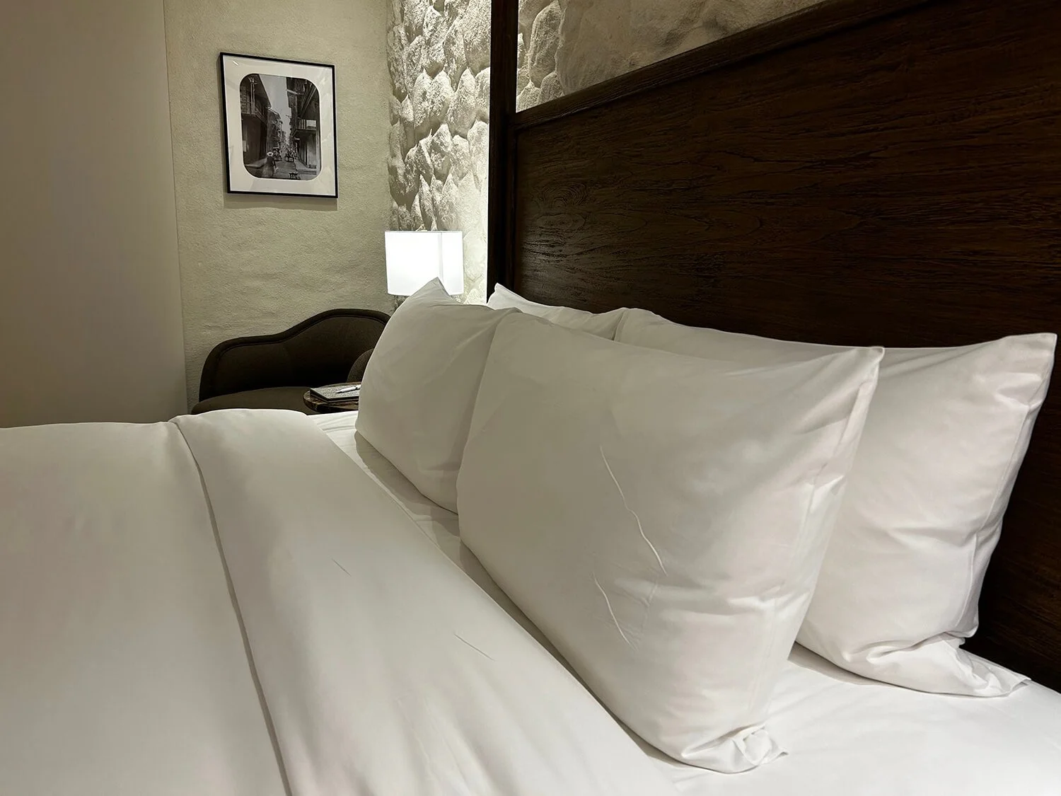 A hotel room with white bedsheets and pillows, a wooden headboard, a side table with a lamp, a black and white framed photograph on the wall, and a small sitting area with a dark chair, a telephone, and a stone accent wall.