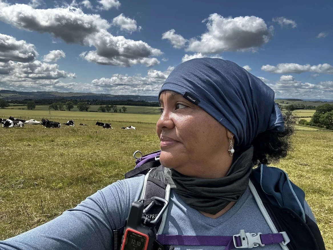 woman hiker looking sideways at cows in background of countryside field blue skies with clouds