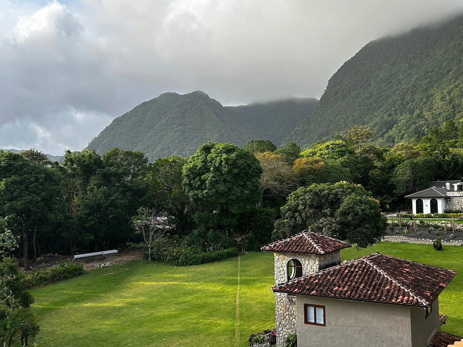 Scenic view of lush green trees and mountains under cloudy sky, with houses and a well-maintained lawn in the foreground.