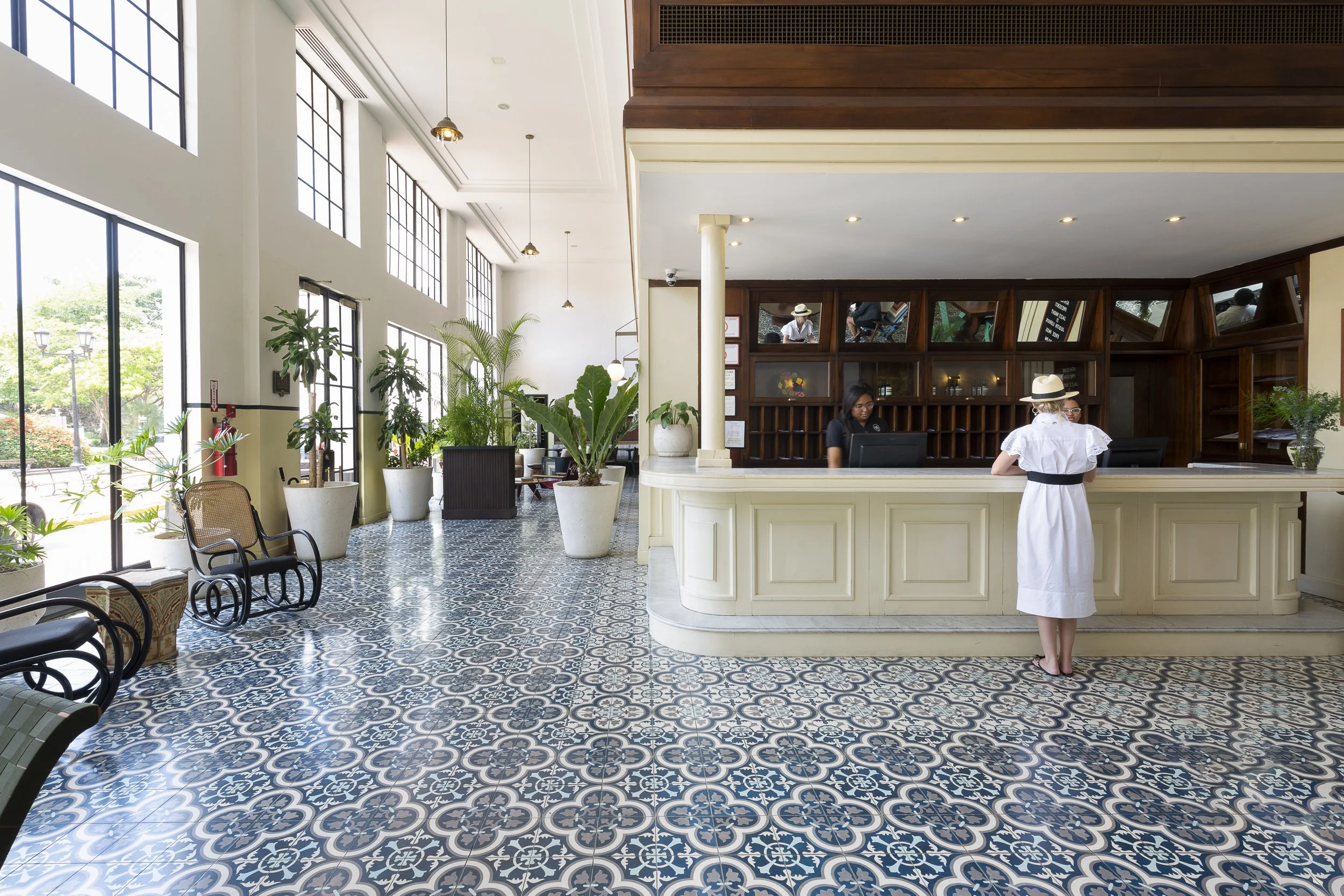 Interior of a bright hotel lobby with patterned tile floor, large windows, green plants, and a reception desk where a woman is checking in with staff. A woman with a hat and white dress stands at the counter.