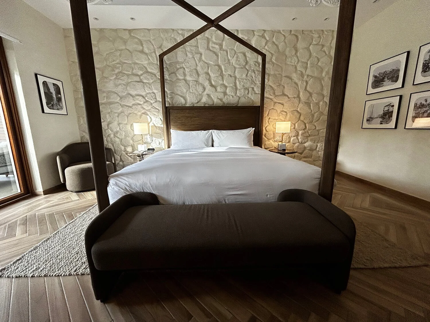 A bedroom with a four-poster bed, white bedding, stone accent wall, two nightstands with lamps, a chair, artwork on the walls, and a glass door leading to a balcony.