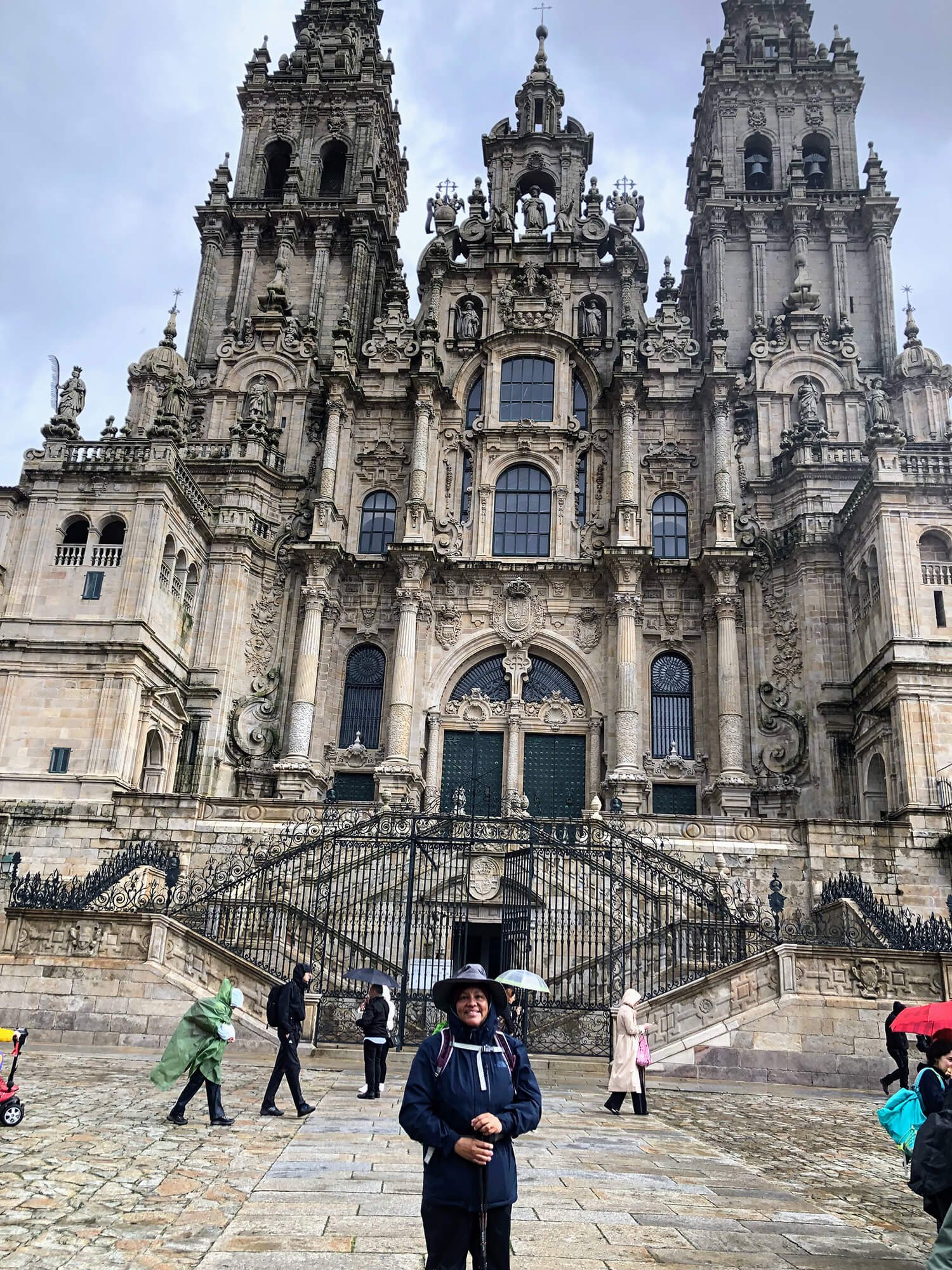 A woman standing in front of a large, ornate building with two towers, some people walking with umbrellas, and cobblestone pavement.