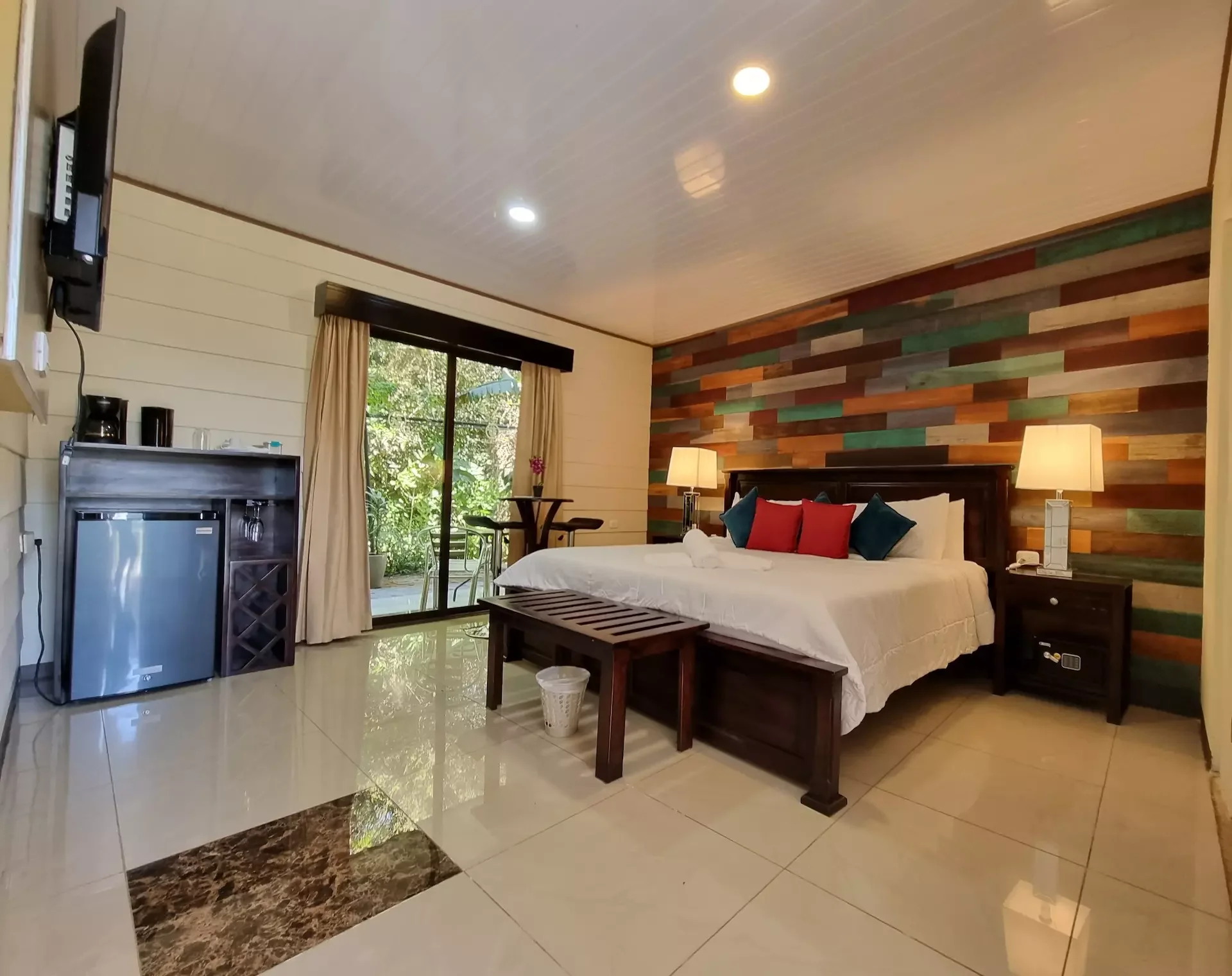 A bedroom with a large bed decorated with pillows, bedside lamps, a wooden bench at the foot of the bed, a small refrigerator, a TV, and a sliding glass door leading to a balcony with trees outside.