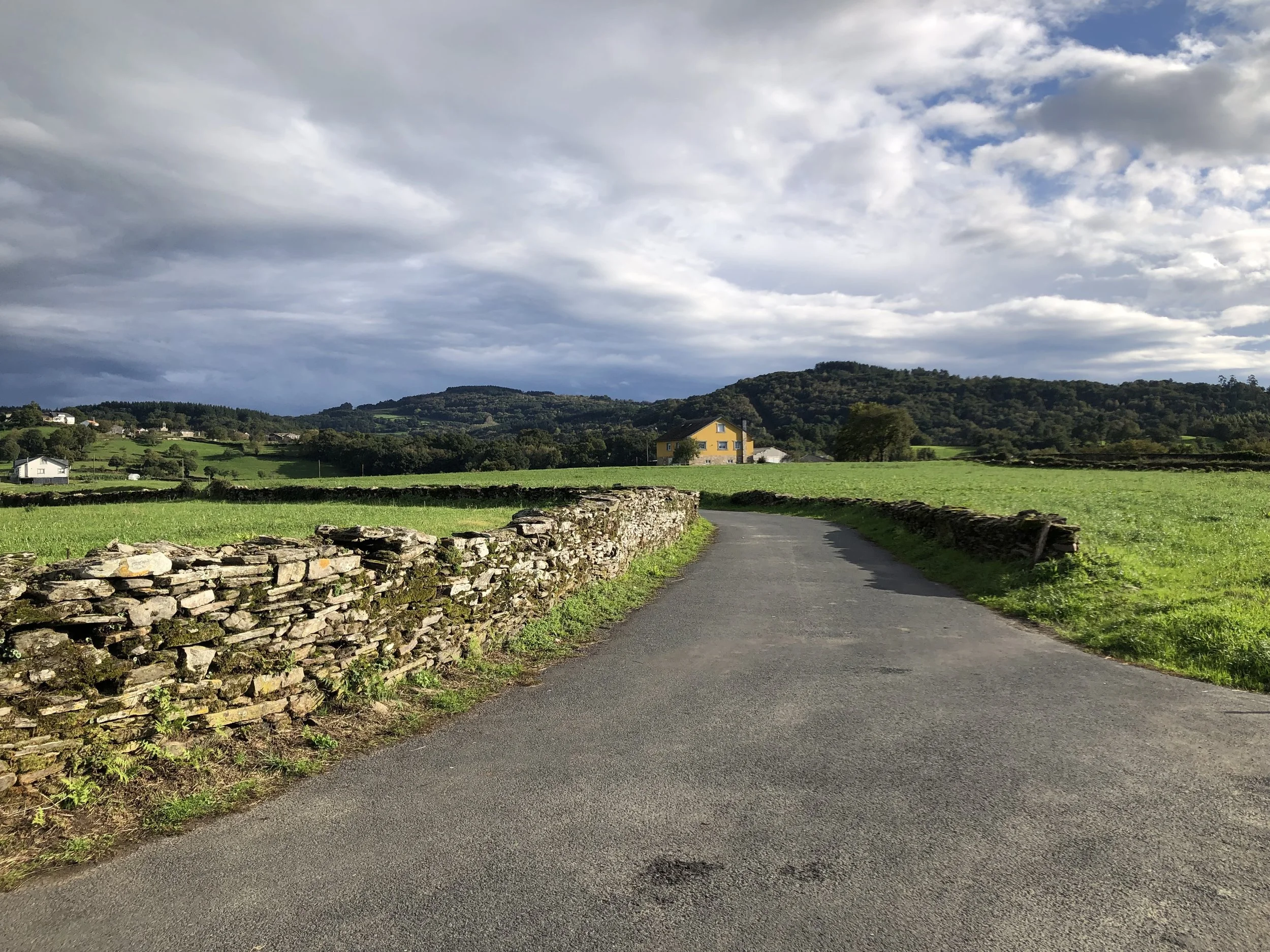 A winding paved country road with a stone wall on the left and green grassy fields on both sides, leading towards rolling hills with scattered houses under a cloudy sky.