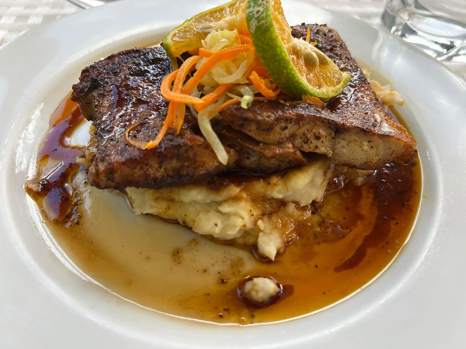 A plate of grilled chicken with mashed potatoes, topped with shredded vegetables and lime wedge, served with gravy and a glass of water.