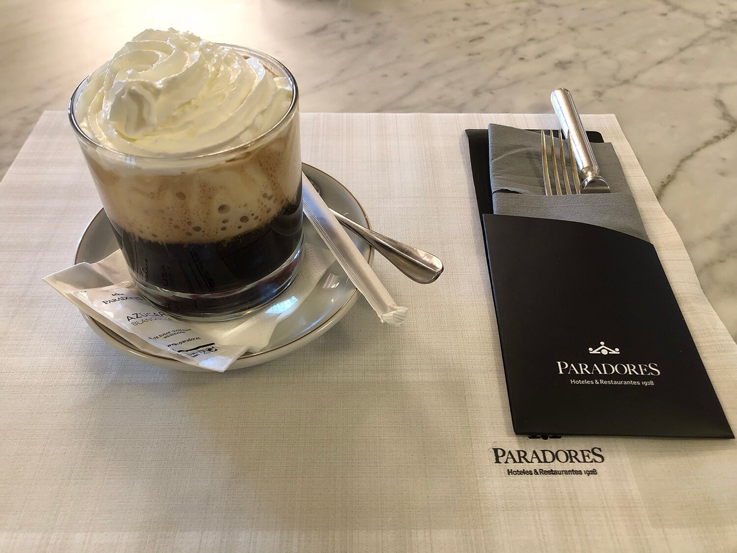 A glass of black coffee topped with whipped cream on a saucer, with a sugar packet and stir stick, placed on a white placemat. To the right, folded napkin with a fork and knife, on a black notepad with the logo and name 'Paradores' and the text 'Hote