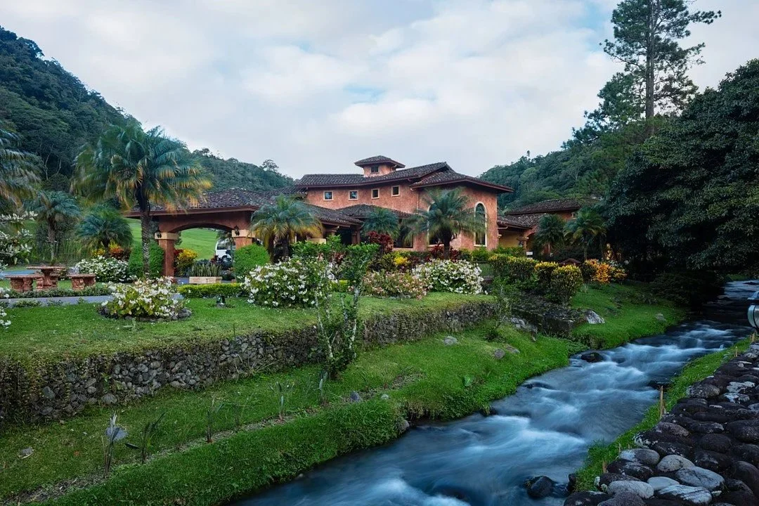 A scenic view of a large house with a garden, palm trees, and flowers beside a flowing creek with rocky banks, surrounded by green hills and trees.