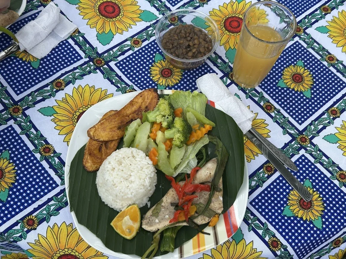 A plate of food with rice, fried fish, salad with lettuce and carrots, grilled chicken with vegetables, lemon wedge, and banana leaf, set on a table with a sunflower-patterned tablecloth. There is a bowl of beans, a glass of orange juice, and a fork 