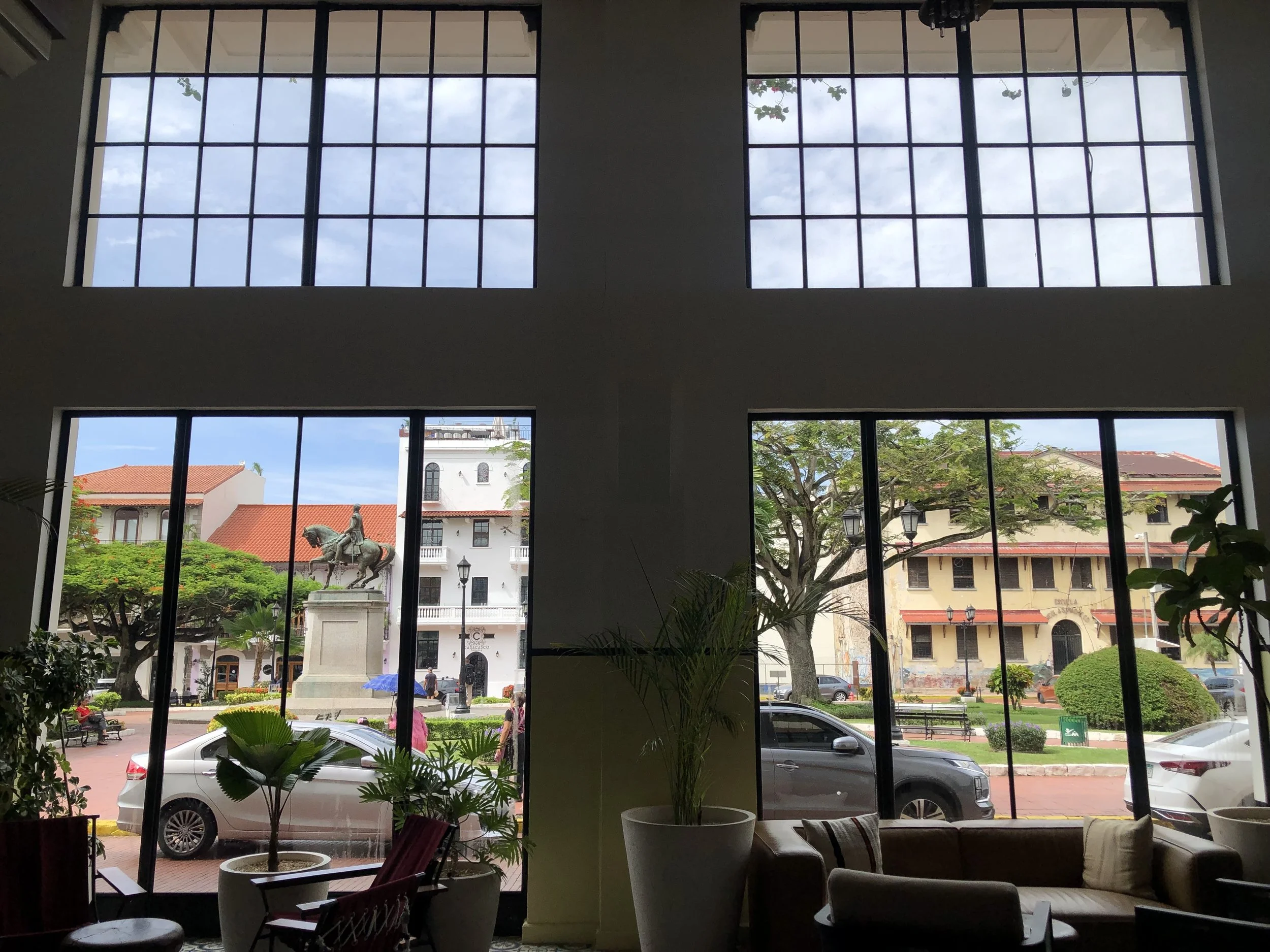 Interior view of a building with large windows showing an outdoor scene with trees, a statue, people sitting on benches, and shops across the street, with some cars parked and driving by.