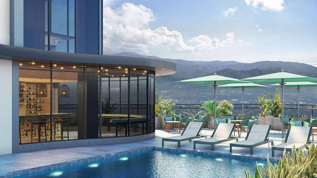 Rooftop pool area with lounging chairs, large umbrellas, potted plants, and a view of distant mountains or hills under a partly cloudy sky.