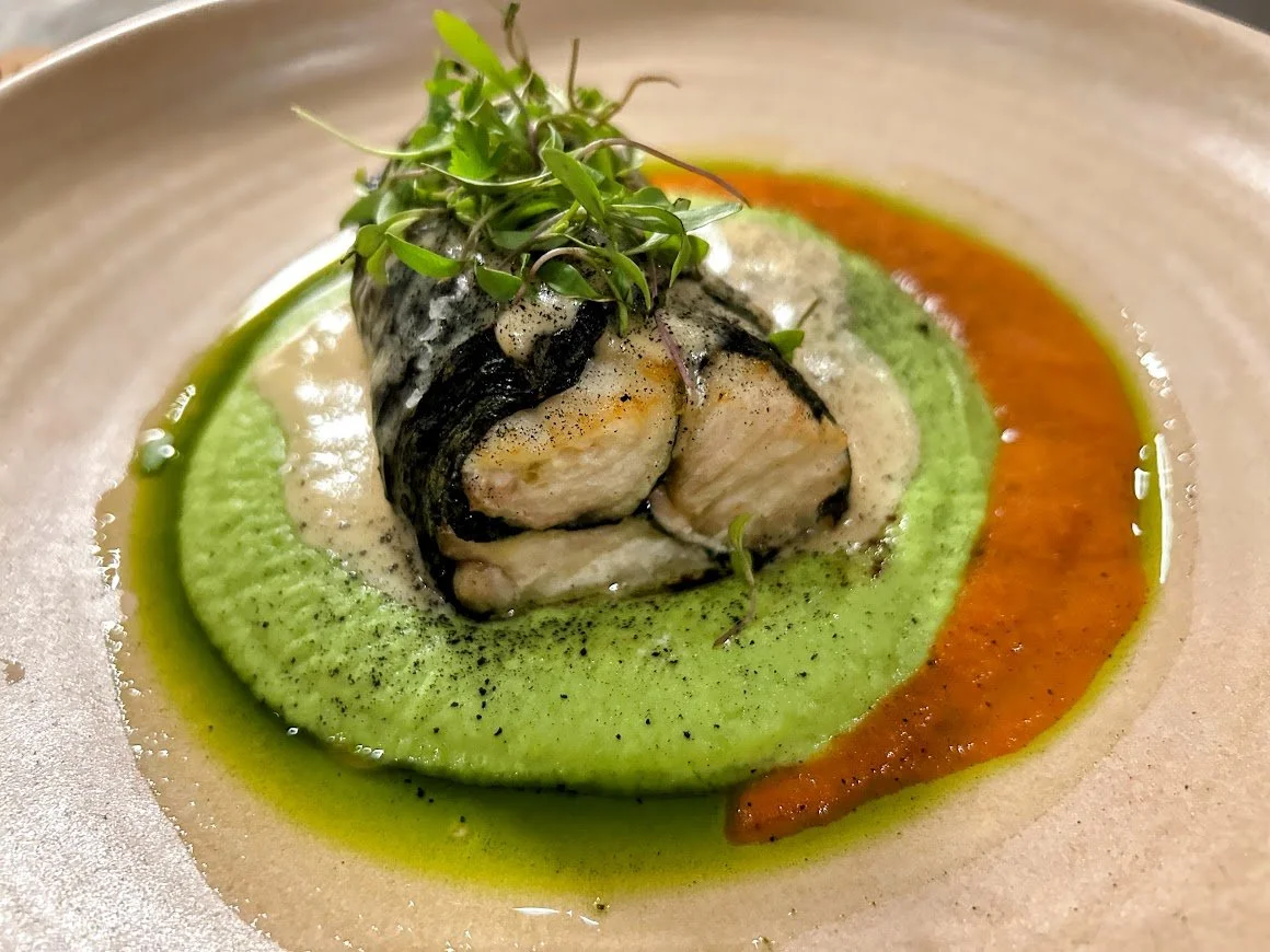 Seared fish fillet with blackened skin, topped with greens, served on a green puree, with orange sauce and foam, garnished with microgreens.