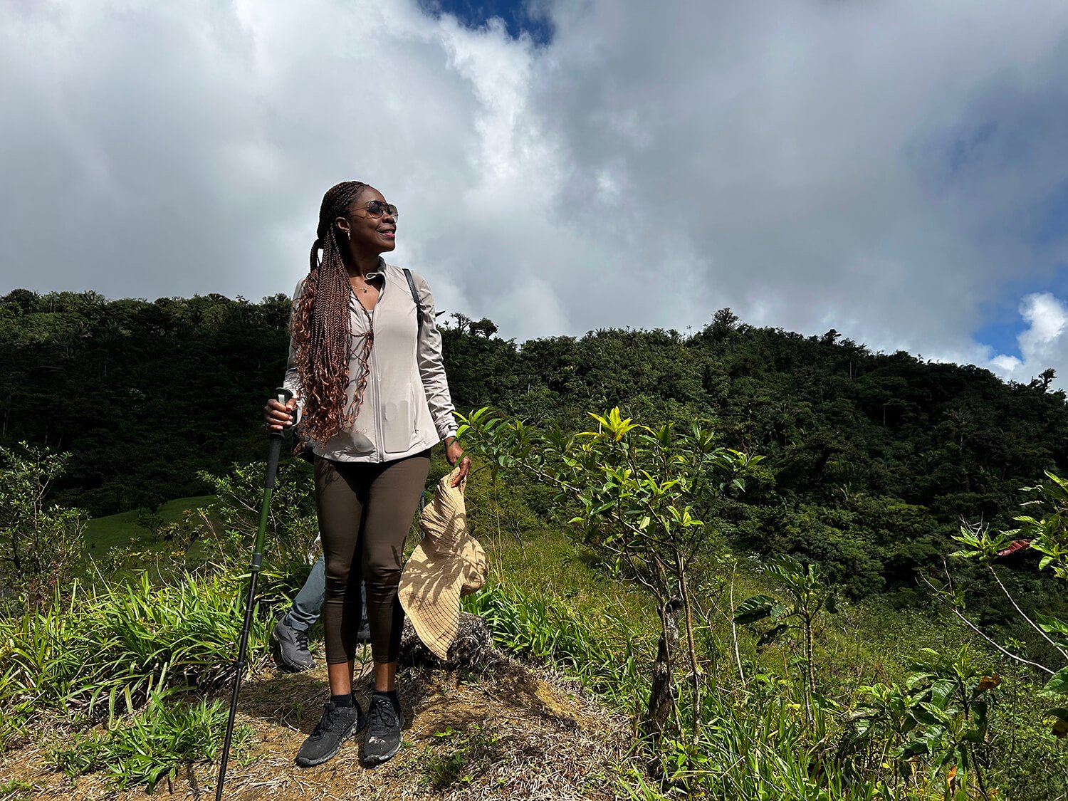 A woman with long braided hair wearing sunglasses, a light jacket, and hiking shoes, holding a hiking pole and a straw hat, standing on a trail in a lush green hilly landscape under partly cloudy sky.