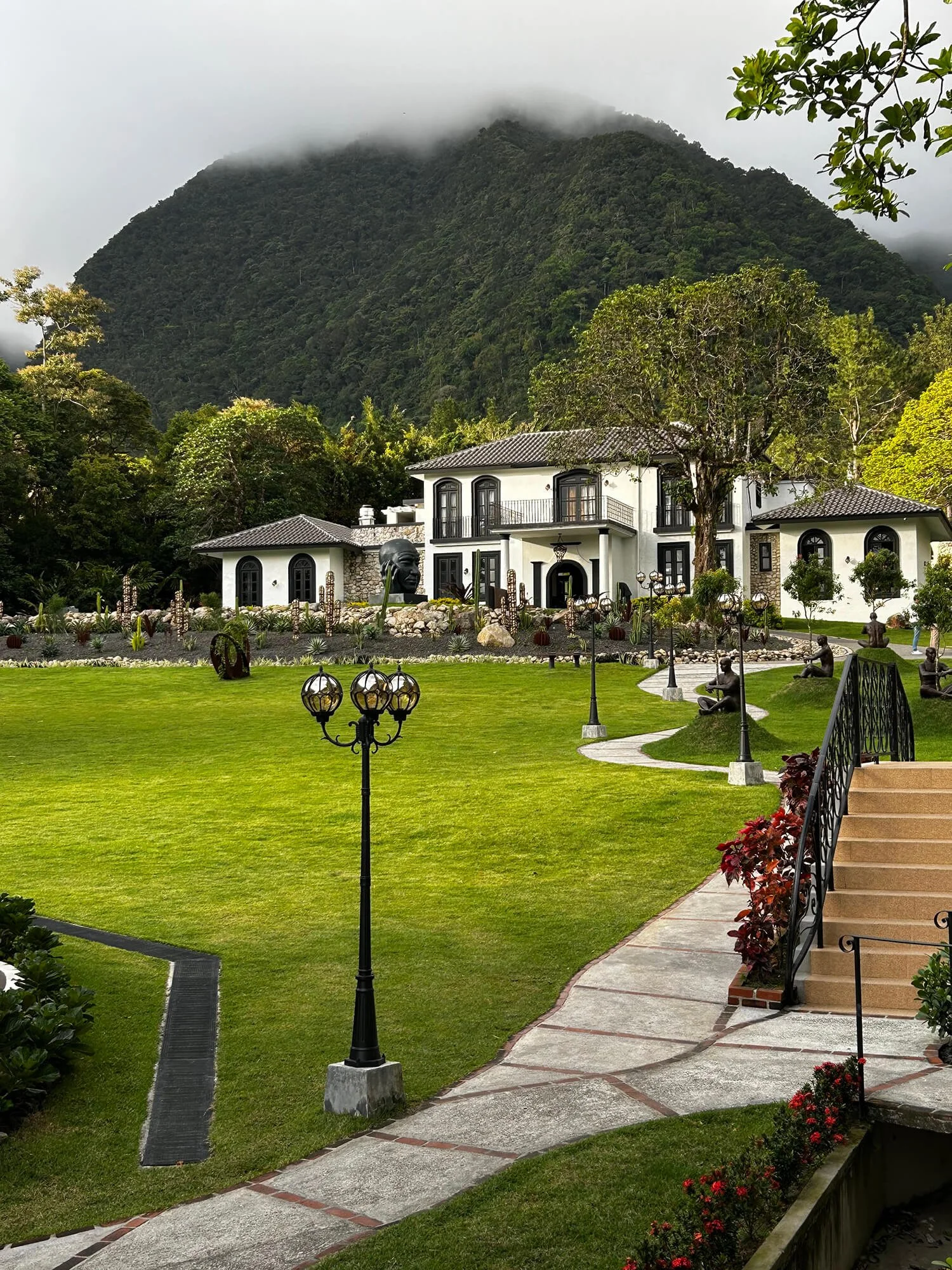 A large white house with black trim and a tiled roof, surrounded by a garden with a manicured lawn, decorative statues, and street lamps. There is a mountain with fog in the background.