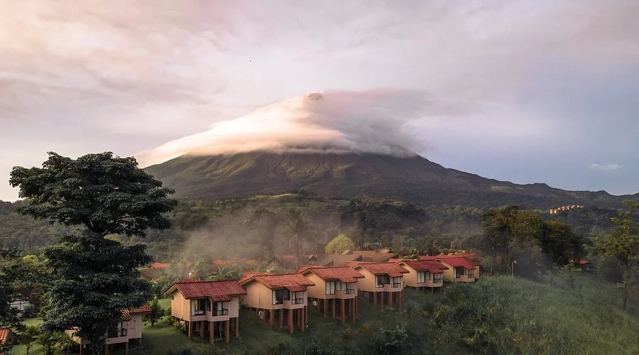 Series of overwater bavilions with red-tiled roofs in lush green landscape with a large mountain in the background, partially covered by clouds.