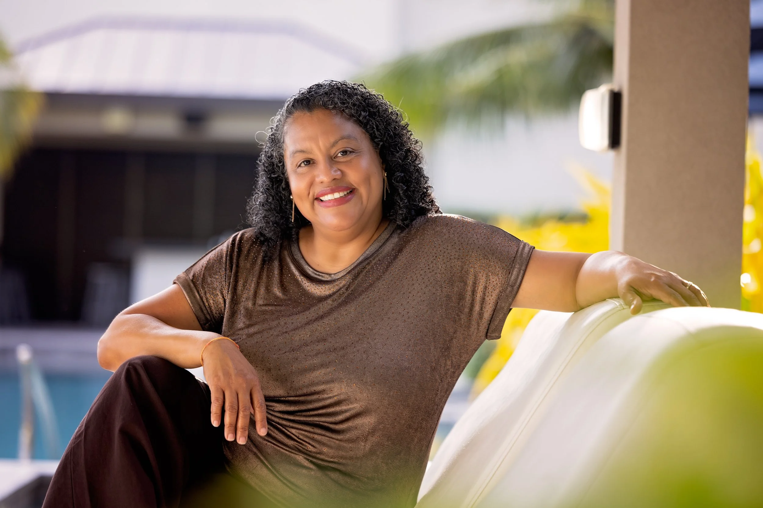 A smiling woman with curly hair, wearing a brown shirt, sitting outdoors next to some large white pumpkins.