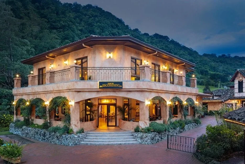 A two-story mountain hotel with a beige stucco exterior, illuminated by exterior lights, surrounded by lush green trees and mountain scenery.