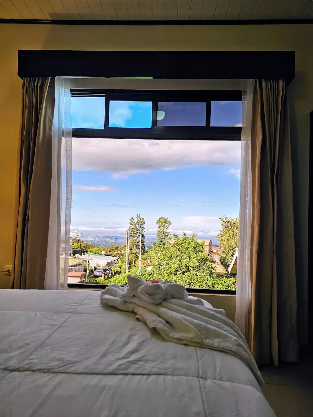 A bedroom with a large window showing a view of trees, houses, and a cloudy sky, with a bed in the foreground covered with a white blanket and folded towels on top.