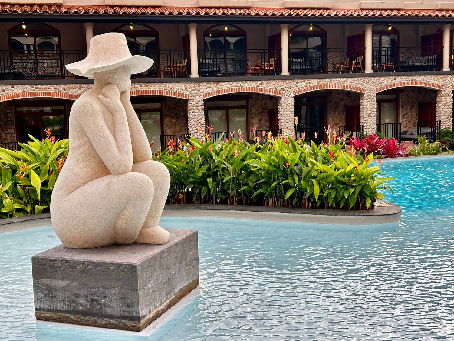 A stone sculpture of a woman wearing a wide-brimmed hat, sitting with her chin rested on her hand, overlooking a swimming pool. Behind her are lush green plants with pink flowers, and a multi-story building with stone and brick facade, balconies, and