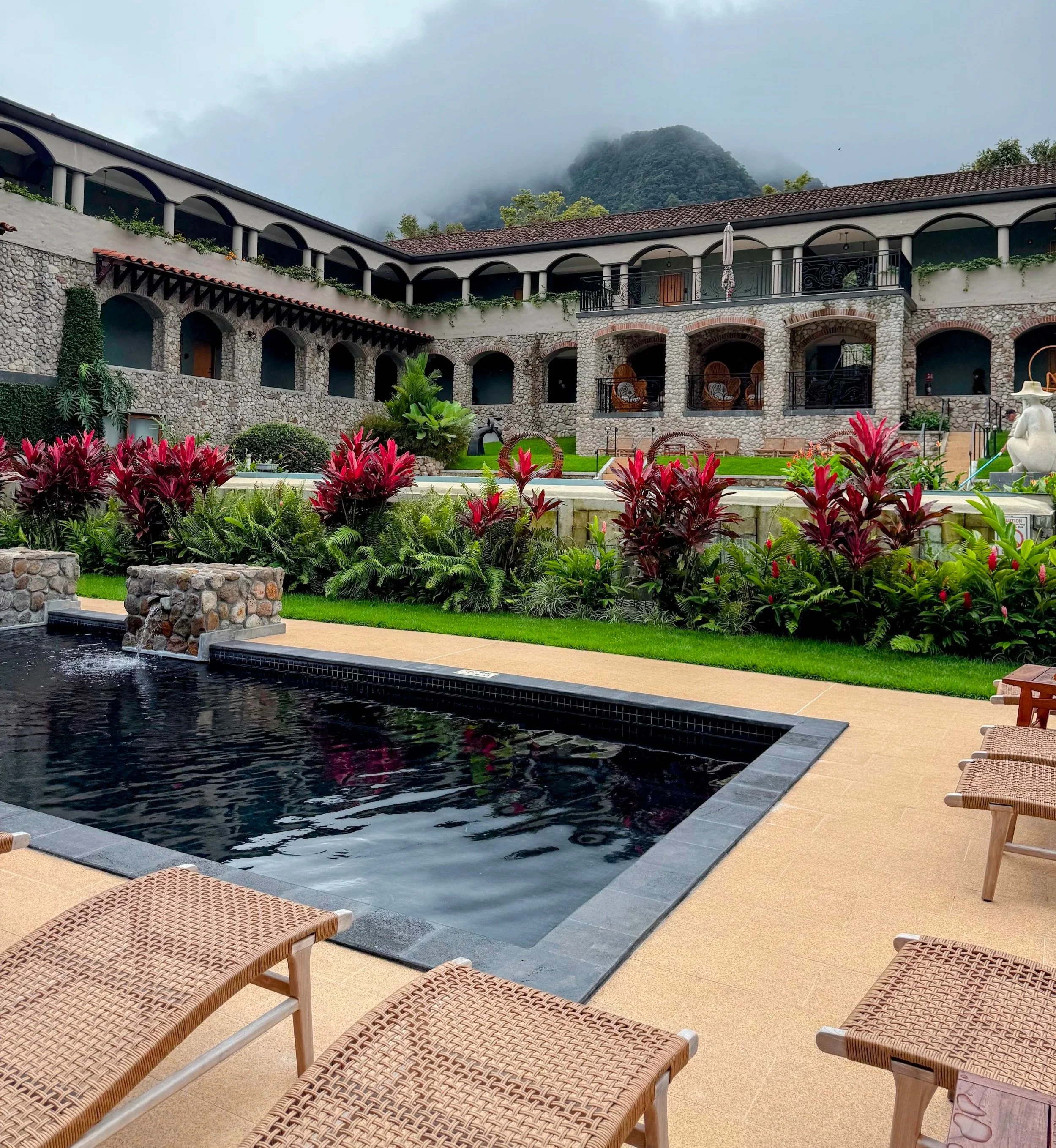 A swimming pool with dark water, surrounded by lounge chairs on beige tiles, with a garden of pink and green plants, and a stone and brick building with arched openings, balconies, and a mountain with fog in the background.