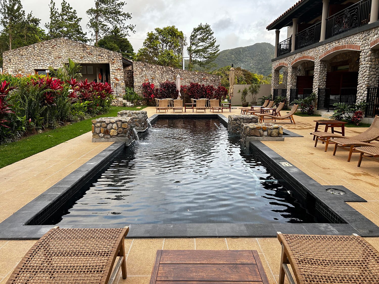 Outdoor pool area with lounge chairs, potted plants, and surrounding landscaping, with stone buildings and mountains in the background.