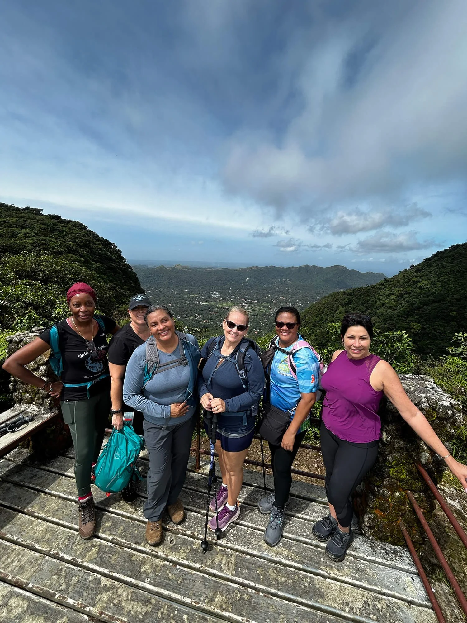 Group of six women hikers standing on a mountain trail with lush green hills and cloudy sky in the background, wearing outdoor gear and smiling.