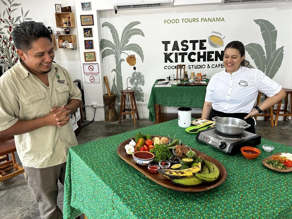 A man and a woman smiling and standing near a table with various fresh vegetables, fruits, and small bowls of sauces or condiments on it inside a restaurant called Taste Kitchen. The woman is preparing food with a portable cooktop, and the man is wat